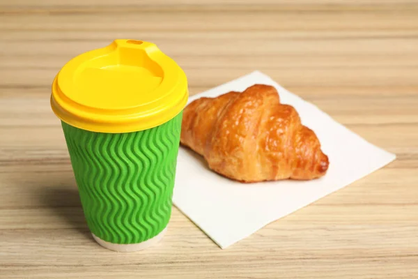Paper cup with coffee and croissant on wooden table