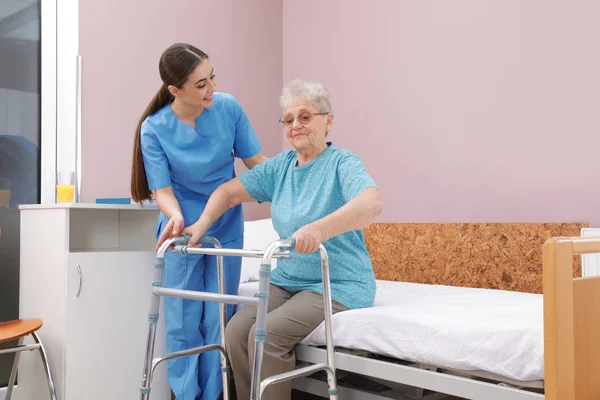 Sjuk sköterska bistå Senior kvinna med Walker att få upp från sängen i sjukhusavdelningar — Stockfoto