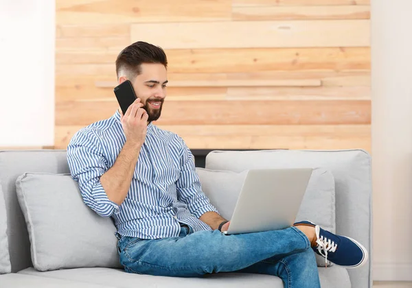 Evde kanepede laptop ile çalışırken telefonda konuşan yakışıklı genç adam — Stok fotoğraf