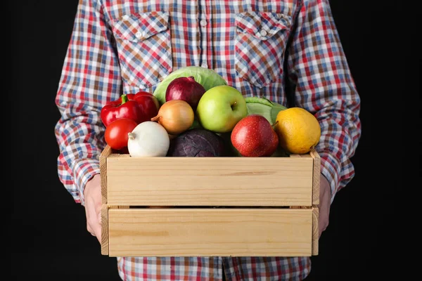 Hombre sosteniendo cajón de madera lleno de verduras frescas y frutas sobre fondo negro, primer plano — Foto de Stock