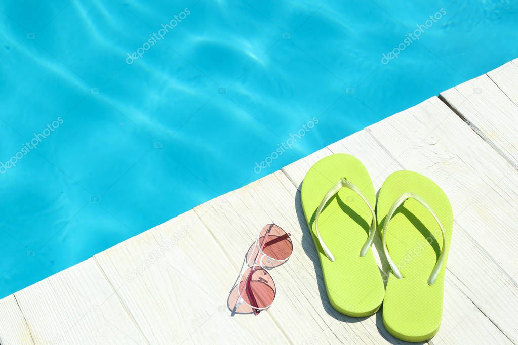 Slippers and sunglasses on wooden deck near swimming pool, space for text. Beach accessories