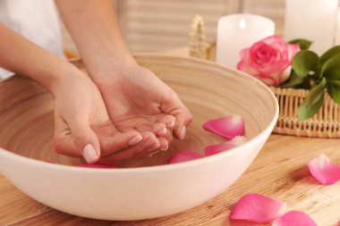 Woman soaking her hands in bowl with water and petals on table, closeup. Spa treatment