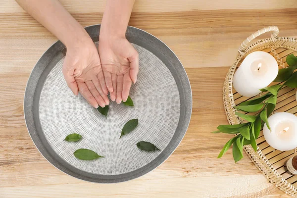 Femme trempant ses mains dans un bol avec de l'eau et des feuilles sur une table en bois, vue sur le dessus. Traitement spa — Photo