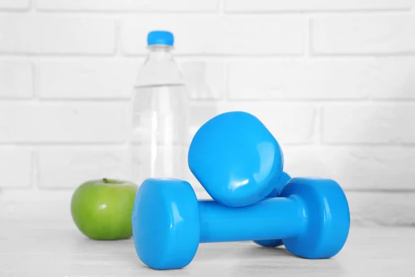 Stylowe hantle, jabłko i butelka wody na stole przed ceglanym murem. Fitness w domu — Zdjęcie stockowe