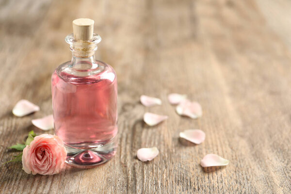 Bottle of rose essential oil and flower on wooden table, space for text