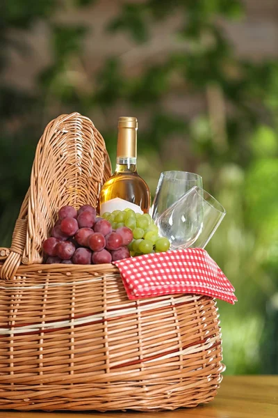 Корзина для пикника с вином, бокалами и виноградом на деревянном столе на размытом фоне, место для текста — стоковое фото