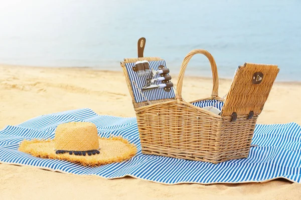 Cesta de vime com piquenique essencial e chapéu de palha no cobertor perto do mar — Fotografia de Stock
