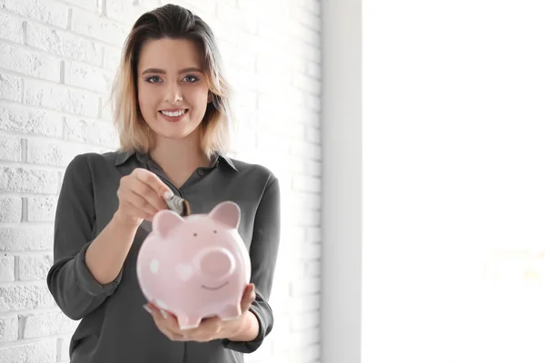 Hermosa mujer poniendo dinero en alcancía cerca de la pared de ladrillo. Espacio para texto — Foto de Stock