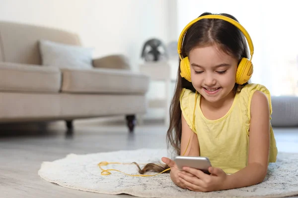 Criança bonito com fones de ouvido e telefone celular no chão dentro de casa. Espaço para texto — Fotografia de Stock