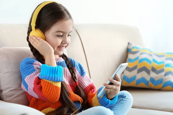 Criança bonito com fones de ouvido e telefone celular no sofá dentro de casa — Fotografia de Stock
