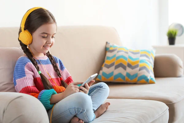 Criança bonito com fones de ouvido e telefone celular no sofá dentro de casa. Espaço para texto — Fotografia de Stock