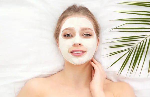 Beautiful woman with clay facial mask and tropical leaf on white fabric, above view — Stock Photo, Image