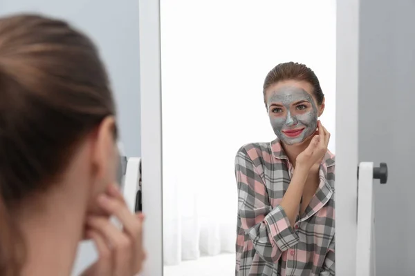 Młoda kobieta z maski oczyszczającej na jej twarzy w pobliżu lustra w łazience. Pielęgnacja skóry — Zdjęcie stockowe