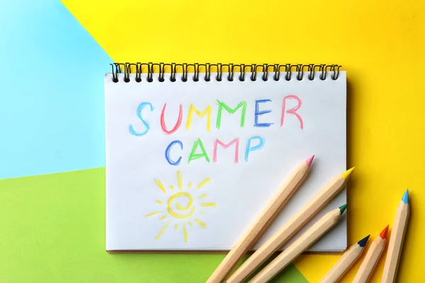 Блокнот з написаним текстом SUMMER CAMP і різними олівцями на кольоровому фоні, вид зверху — стокове фото