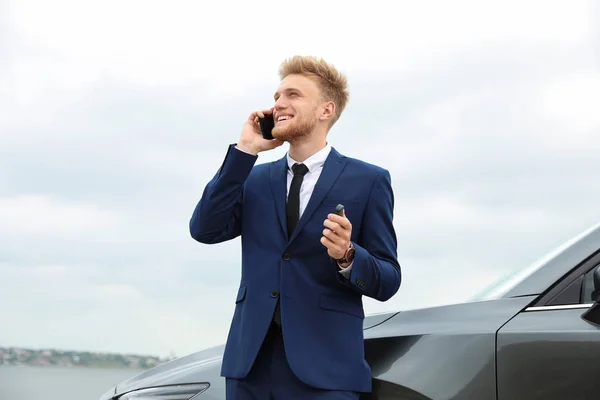 Fiatal üzletember kulccsal beszél telefonon a kocsi mellett a szabadban — Stock Fotó