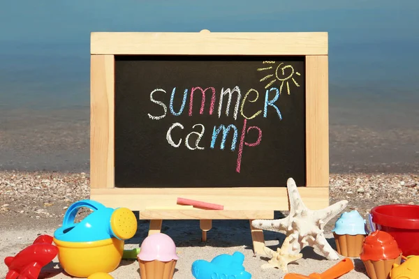 Kleine Kreidetafel mit Text Sommerlager und Strand Spielzeug auf Sand in der Nähe des Flusses — Stockfoto
