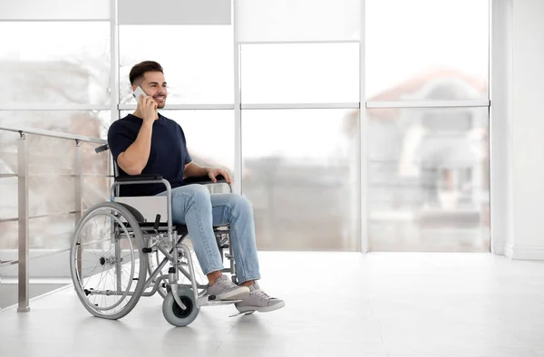 Молодой человек в инвалидной коляске разговаривает по мобильному телефону возле окна в помещении — стоковое фото