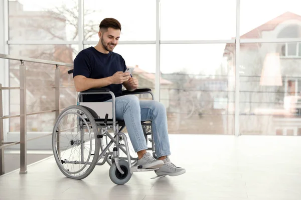 Junger Mann im Rollstuhl benutzt Handy in der Nähe von Fenster — Stockfoto