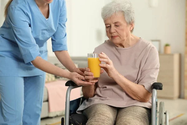 Enfermera dando vaso de jugo a anciana en silla de ruedas en interiores. Asistencia a personas mayores —  Fotos de Stock