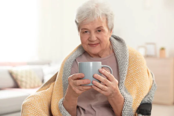 Elderly woman covered with blanket drinking tea at nursing home. Assisting senior people