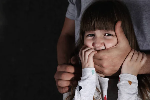 Взрослый мужчина, прикрывающий рот испуганной маленькой девочки на тёмном фоне, место для смс. Ребенок в опасности — стоковое фото
