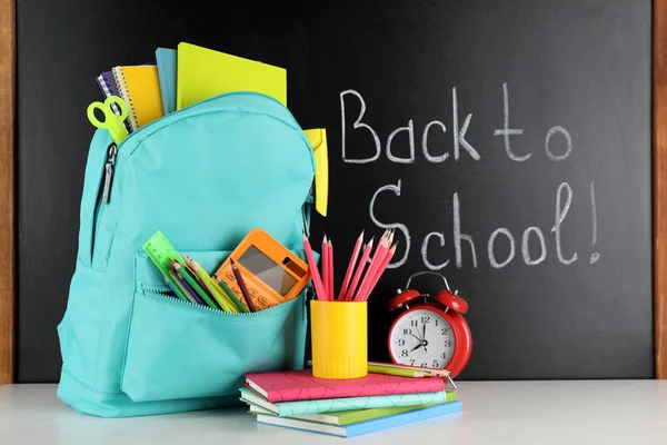 Mochila brillante con papelería escolar en mesa blanca cerca de pizarra negra. Volver a la escuela — Foto de Stock