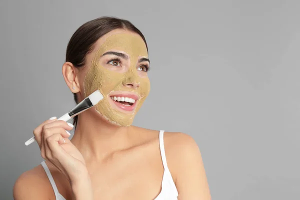 Jonge vrouw het toepassen van klei masker op haar gezicht tegen grijze achtergrond. Ruimte voor tekst — Stockfoto