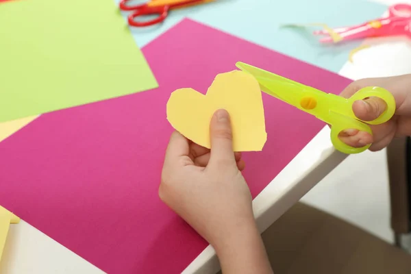 子供はテーブルでプラスチックハサミで紙の心臓を切り取り、クローズアップ。テキスト用のスペース — ストック写真