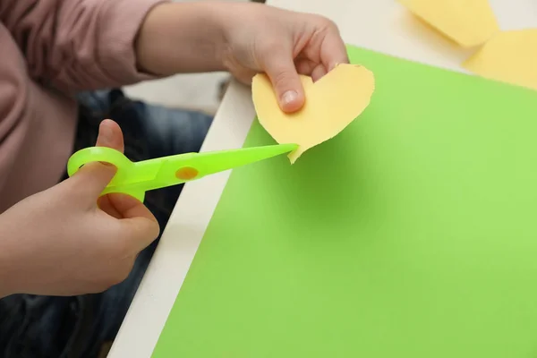 Child cutting out paper heart with plastic scissors at table, closeup. Space for text — Stock Photo, Image