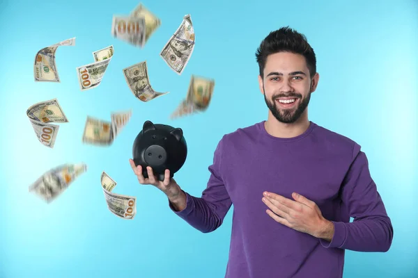 Glücklicher junger Mann mit Sparschwein und fallendem Geld auf blauem Hintergrund — Stockfoto