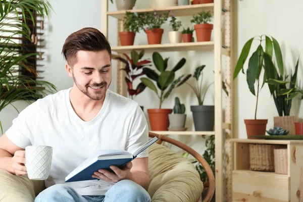 Junger Mann liest Buch im Zimmer mit verschiedenen heimischen Pflanzen — Stockfoto