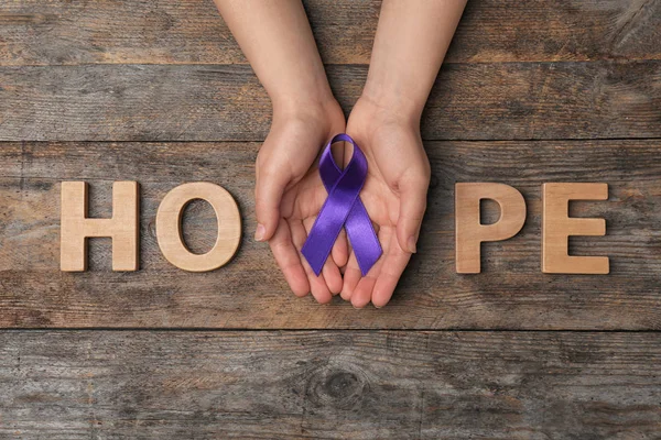 Woman with purple awareness ribbon and word HOPE on wooden background, top view