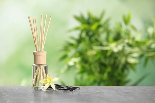 Reed ambientador com flor de baunilha e varas na mesa cinza contra fundo verde desfocado. Espaço para texto — Fotografia de Stock