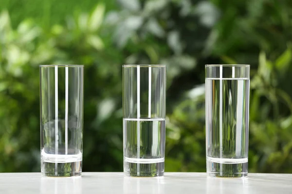 Vaciado, medio y lleno vasos de agua sobre una mesa blanca sobre fondo borroso — Foto de Stock