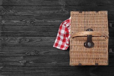 Siyah ahşap masa üzerinde damalı masa örtüsü ve kapalı hasır piknik sepeti, üst görünümü. Metin için alan