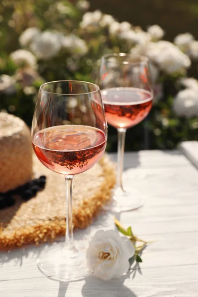 Glasses of rose wine, straw hat and beautiful flower on white wooden table outdoors