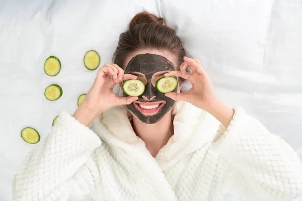 Młoda kobieta z maską twarzy i plastry ogórka na łóżku, widok z góry — Zdjęcie stockowe