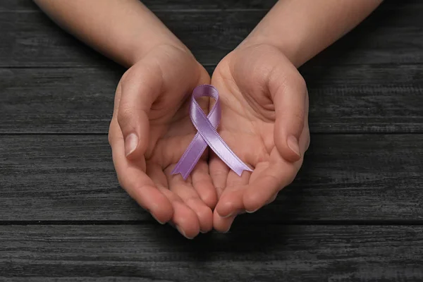 Woman holding purple awareness ribbon on black wooden background, closeup
