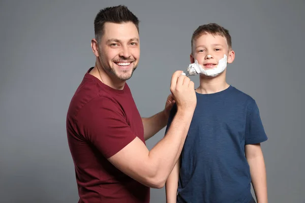Tata stosujące piankę do golenia na twarzy syna, szare tło — Zdjęcie stockowe