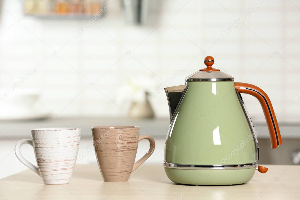 Stylish electrical kettle and cups on table against blurred room interior. Space for text