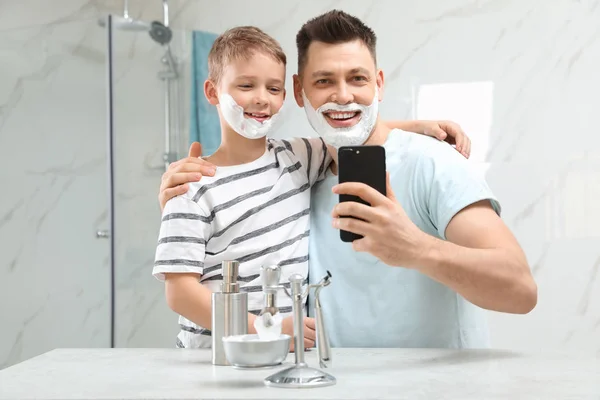 Сын и папа с пеной для бритья на лицах делают селфи в ванной — стоковое фото