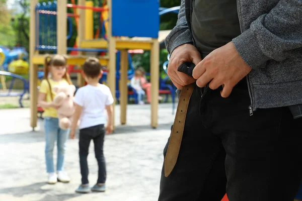 Un uomo adulto sospetto che si toglie i pantaloni al parco giochi con bambini piccoli, spazio per messaggi. Bambino in pericolo — Foto Stock