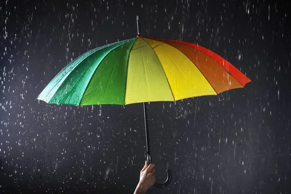 Mulher segurando guarda-chuva brilhante sob chuva no fundo escuro, close-up — Fotografia de Stock