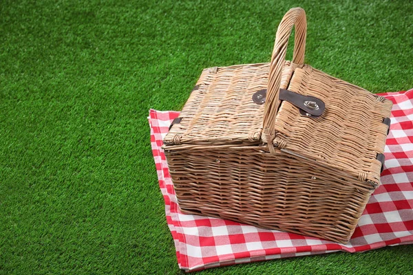 Checkered tablecloth and closed wicker picnic basket on green grass, space for text — Stock Photo, Image