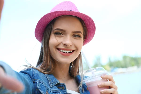 Happy young woman with drink taking selfie on riverside