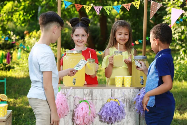 Cute little girls selling natural lemonade to boys in park. Summer refreshing drink — Stock Photo, Image