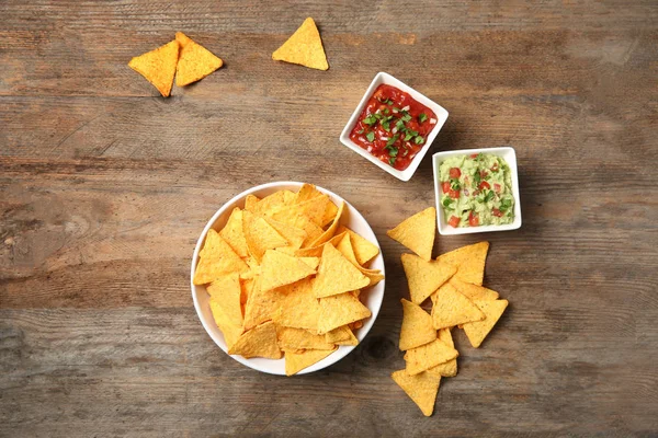 Bowl of delicious mexican nachos chips and sauces on wooden table, flat lay — Stock Photo, Image