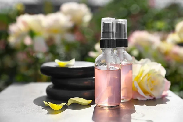 Piedras de spa, botellas de tóner con aceite esencial y rosa en la mesa en el jardín floreciente. Armonía y zen — Foto de Stock