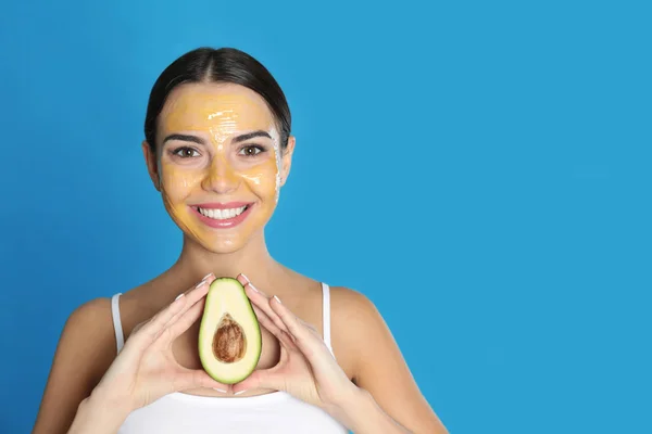 Jonge vrouw met reinigende masker op haar gezicht houden van avocado tegen kleur achtergrond, ruimte voor tekst. Huidverzorging — Stockfoto