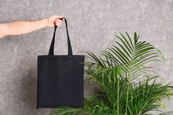 Joven sosteniendo bolsa ecológica en planta de palma interior cerca de pared gris —  Fotos de Stock